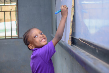 African girl child, pupil or student sitting down and writing in a classroom while studying for...