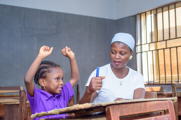African Nigerian mother or teacher sitting together with her girl child in a classroom, helping her...