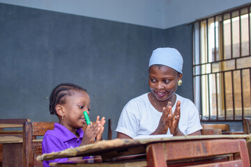 African Nigerian mother or teacher sitting together with her girl child in a classroom, helping her...