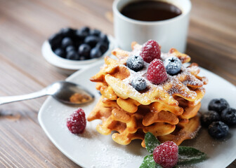 Homemade Belgian waffles served with fresh berries on white plate with a cup of coffee over wooden background, close up.