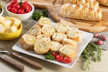 Garlic bread in white square plate on the table with cheese, rosemary, olives and cherry tomatoes.