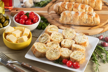Garlic bread in white square plate on the table with cheese, rosemary, olives and cherry tomatoes.