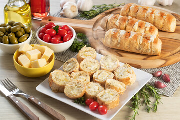 Garlic bread in white square plate on the table with cheese, rosemary, olives and cherry tomatoes.