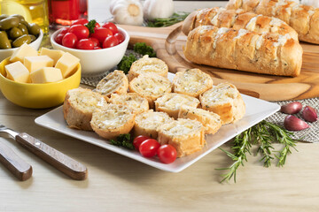 Garlic bread in white square plate on the table with cheese, rosemary, olives and cherry tomatoes.