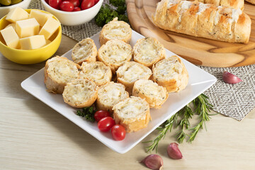 Garlic bread in white square plate on the table with cheese, rosemary, olives and cherry tomatoes.