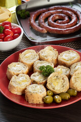 Garlic bread on a red plate on the barbecue table with sausage, cheese, rosemary, olives and cherry tomatoes.