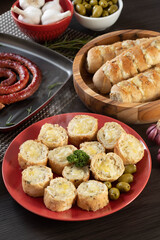 Garlic bread on a red plate on the barbecue table with sausage, cheese, rosemary, olives and cherry tomatoes.