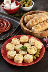 Garlic bread on a red plate on the barbecue table with sausage, cheese, rosemary, olives and cherry tomatoes.