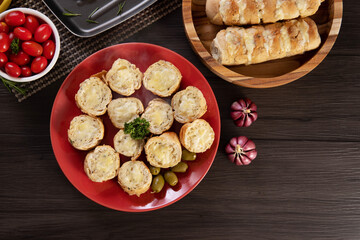 Garlic bread on a red plate on the barbecue table with sausage, cheese, rosemary, olives and cherry tomatoes. Top view.