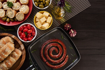 Sausage and garlic bread on a red plate on the barbecue table with appetizers, cheese, rosemary, olives and cherry tomatoes. Top view.