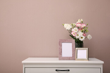Empty photo frames and beautiful bouquet on white chest of drawers near pink wall, space for text