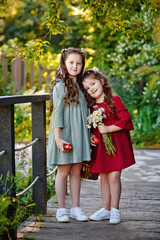 Fototapeta premium Portrait of two girls in a red and green dress, smiling outdoors, enjoying a warm sunny summer day. Sisters with apples and flowers.