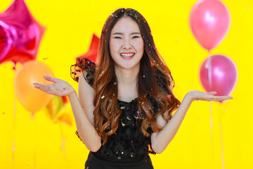 Asian young pretty happy girl in black dress stand smiling surprised excited under throwing glossy paper confetti in front pink star shape balloons on yellow background at Birthday party celebration