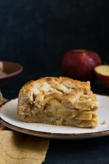 Slice of homemade apple pie with dark background.  Apples and cloth on the background. Close up. Dark mood, rustic, fresh.