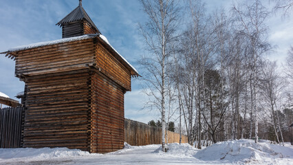 The old watchtower is made of unpainted logs. There is a board fence around the city. There is snow on the ground. Bare trunks and branches of birch trees against the blue sky. Winter day. Siberia