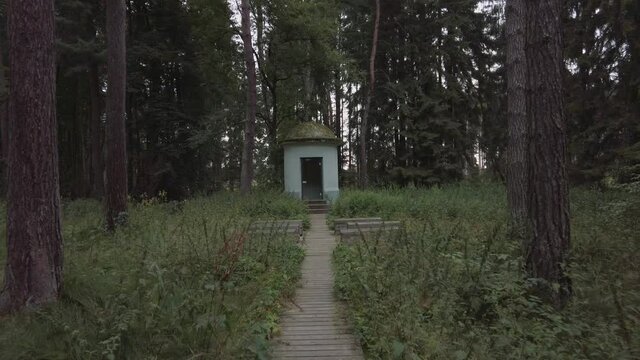 Walking  along a  wooden walkway though a pine forest  slowly approaching a small building with wooden benches in front.