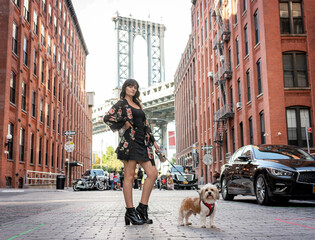 a woman posing with her small mixed breed dog in front of manhattan bridge at dumbo, brooklyn