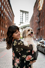 a woman posing with her small mixed breed dog in front of manhattan bridge at dumbo, brooklyn