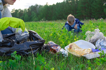 Female hands in yellow rubber gloves collect plastic trash in a black bag outdoors. In the...