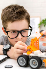 Close-up of a clever boy in eyeglasses builds and programs a robot car at home. The child is learning coding and programming. Robotics, Science, Mathematics, Engineering, Technology. STEM education.
