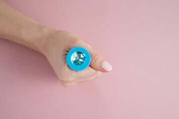 A woman is holding a blue anal plug with a crystal on a pink background. Adult toy for alternative sex