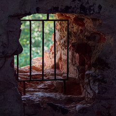 A window with a lattice in a thick brick red stone wall of a medieval fortress: light darkness stones prison lack of freedom.