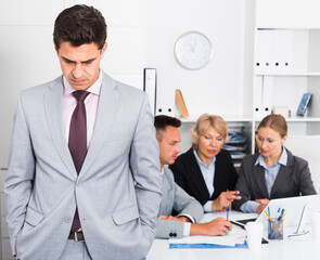 Unhappy and tired man standing at office on background with working colleagues