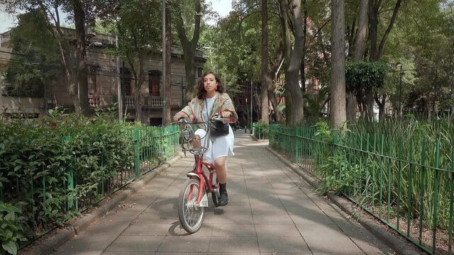 Beautiful young hispanic brunette woman with curly hair smiling and looking at the camera while riding a bike on an urban park