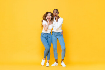 Full length portrait of shocked interracial millennial woman friends gasping with hands cupping mouths in isolated studio yellow color background