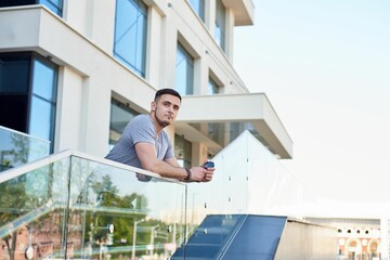 athletic handsome young caucasian brunette guy in a gray t-shirt leaned on a glass railing against the background of a building