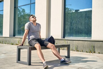 a handsome young Caucasian man with dark hair in sunglasses sits on a bench near the building,...