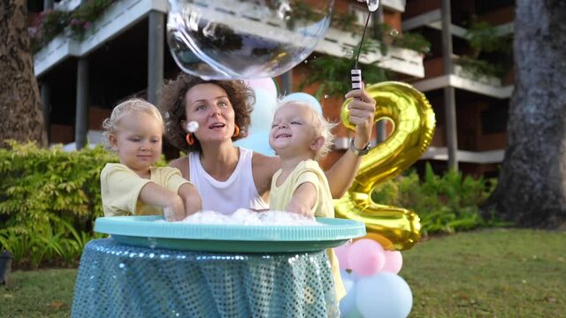 Young mom makes foam bubbles with her twin toddlers at their second birthday party