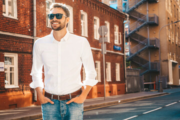 Fototapeta premium Portrait of handsome confident stylish hipster lambersexual model.Modern man dressed in white shirt. Fashion male posing in the street background near skyscrapers in sunglasses