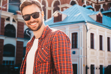 Handsome smiling stylish hipster lambersexual model.Modern man dressed in red checkered shirt. Fashion male posing near skyscraper on the street background in sunglasses. Outdoors at sunset