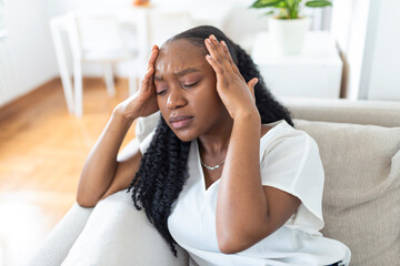 Portrait of a young black girl sitting on the couch at home with a headache and pain. Beautiful woman suffering from chronic daily headaches. Sad woman holding her head because sinus pain