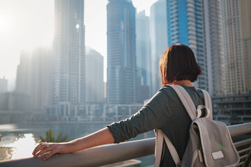 Fototapeta premium Happy young female traveler in the big city of Dubai, famous place Dubai marina. Luxury and comfortable tourism season in United Arab Emirates. Back or rear view of young woman in dress and backpack