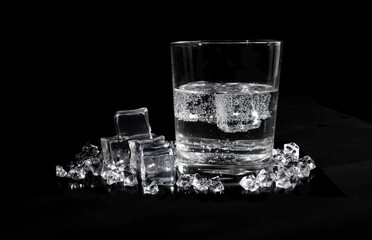 A transparent glass beaker with a colorless liquid ice cubes floating in it and scattered on the black surface on which the glass stands