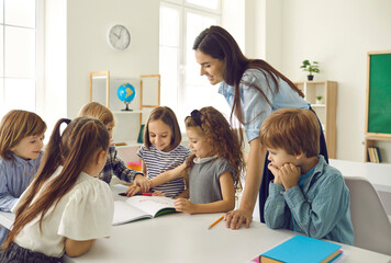 Group of children looking at picture that little girl is showing. Happy school teacher and students have interesting classes, learn, discuss new things, work on creative projects in modern classroom - obrazy, fototapety, plakaty