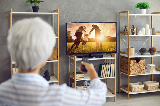 Senior woman watching photo collection of her life memories, family, children and grandchildren, or movie, series or serial on television channel on widescreen TV at home. Back rear view from behind