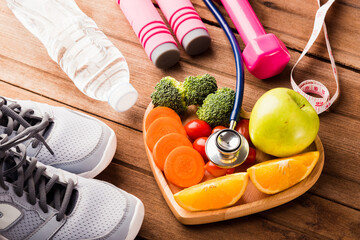 Top view of fresh fruits and vegetables in heart plate wood (apple, carrot, tomato, orange, broccoli) and sports equipment and doctor stethoscope on wooden table, Healthy lifestyle diet food concept - Powered by Adobe