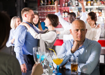 Troubled man sitting alone at office party in nightclub on background with cheerful workmates