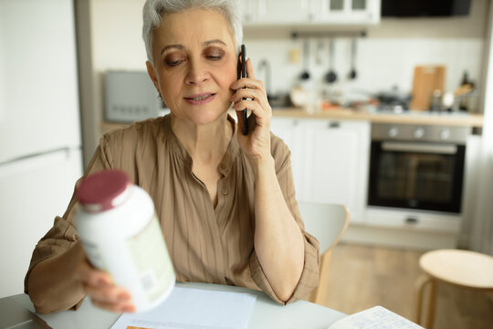 Horizontal Image Of Mature Healthy-looking Senior Lady Sitting At Kitchen Table With Bottle Of Supplements, Calling Her Doctor Or Nutritionist For Detailed Information Upon Vitamin Prescription