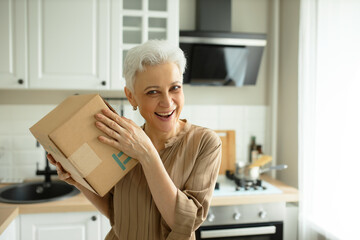 Fototapeta na wymiar Horizontal image of cheerful female of sixty standing in kitchen and shaking carton box in hand, trying to guess what's inside parcel, with happy, delighted face expression. People and online services