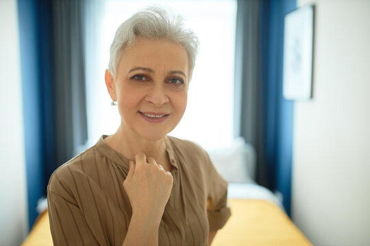 Head shot of mature female blogger with wrinkled skin wearing natural makeup, posing against blurred background of modern bedroom interior, looking at camera with joyful face expression