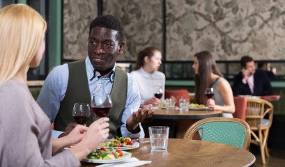 Confident African American businessman with female partner having dinner and drinking red wine at restaurant