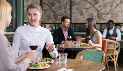Fototapeta na wymiar Cheerful young woman with female friend enjoying dinner and drinking red wine in restaurant
