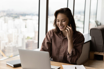 Happy businesswoman talking on smartphone at workplace with laptop. Manager giving telephone consultation to client, speaking, smiling. Secretary answering mobile phone call in office