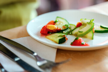 White plate with salad of cucumbers, tomatoes and greens. Next to cutlery.