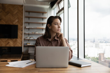 Thoughtful business owner woman looking out of window at city, sitting at workplace with laptop,...