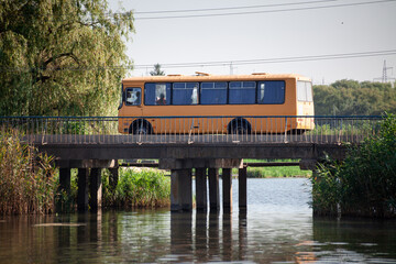Orange bus on concrete bridge over a small river with green reeds. Road bridge with gray concrete columns.